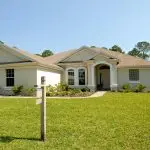 Beautiful suburban house with lush lawn, showcasing a for sale sign under a clear blue sky.