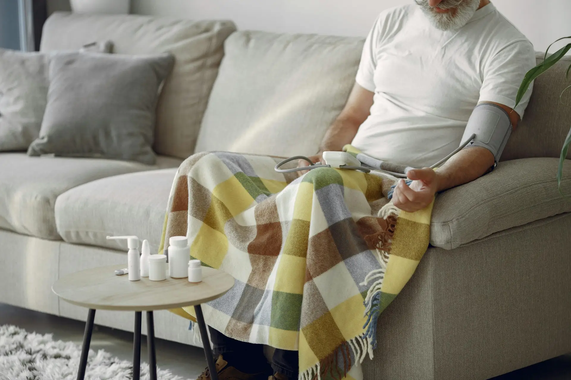 Elderly man monitoring blood pressure on a cozy couch at home, surrounded by medication.