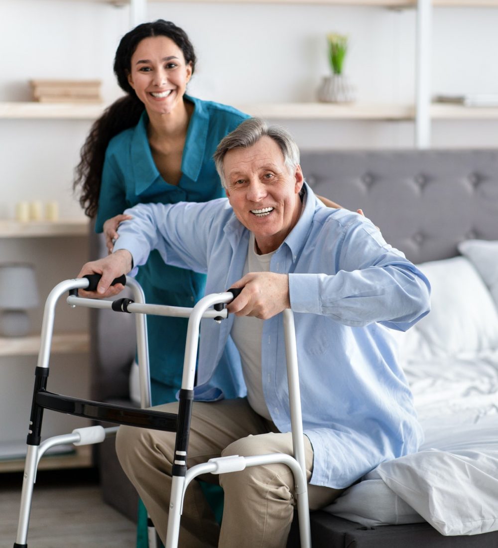 female-nurse-helping-elderly-male-with-walking-frame-stand-up-from-bed-at-home-professional-care.jpg